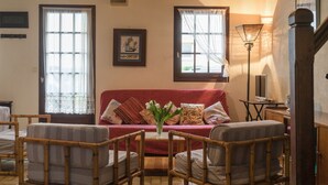 Un salon chaleureux avec un canapé rouge, deux fauteuils en osier, une petite table sur laquelle est posé un vase de fleurs et une lampe.