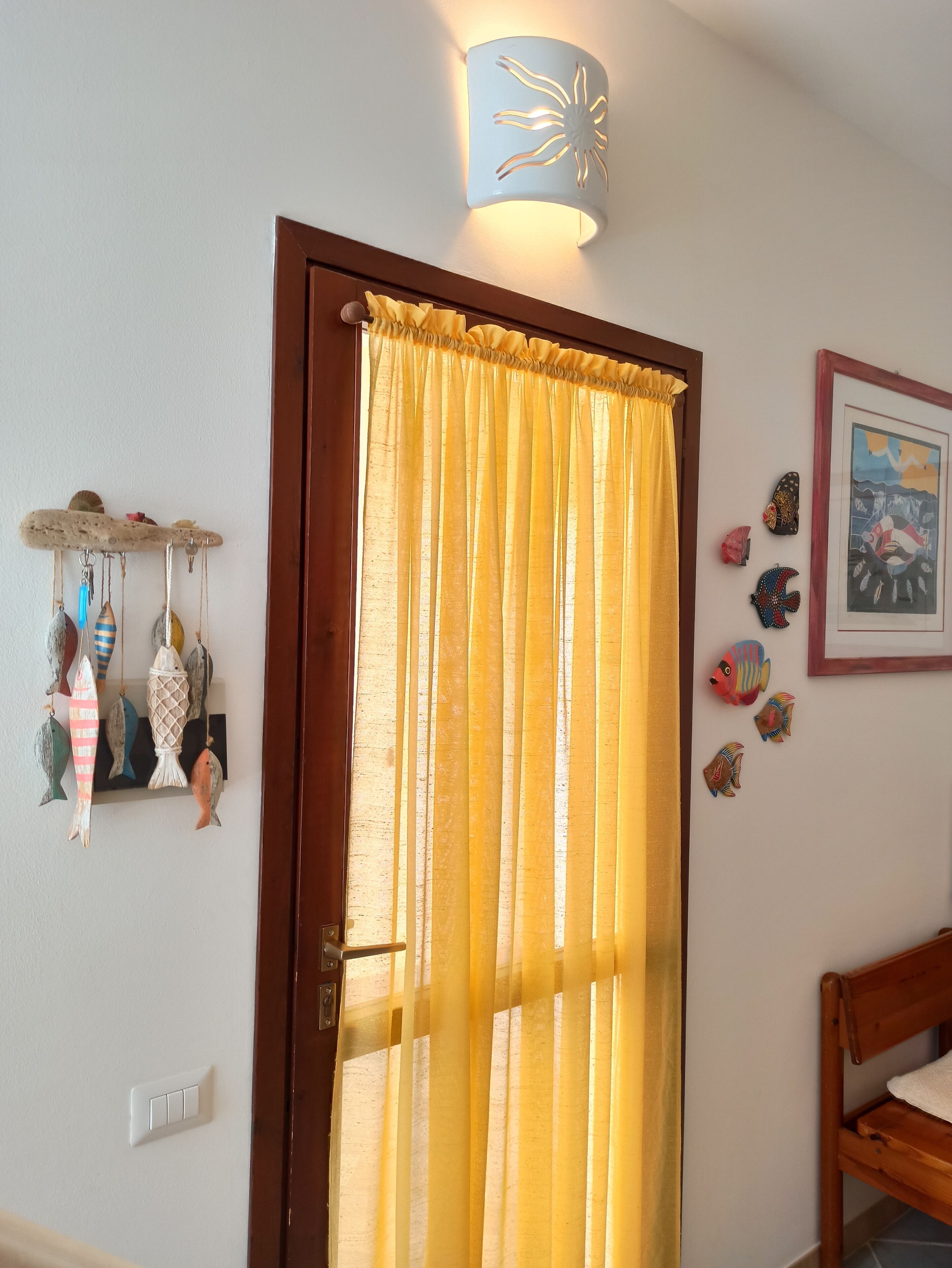 Une pièce avec une porte en bois, des rideaux jaunes, un luminaire fixé au mur et une étagère ornée de poissons et de coquillages décoratifs.