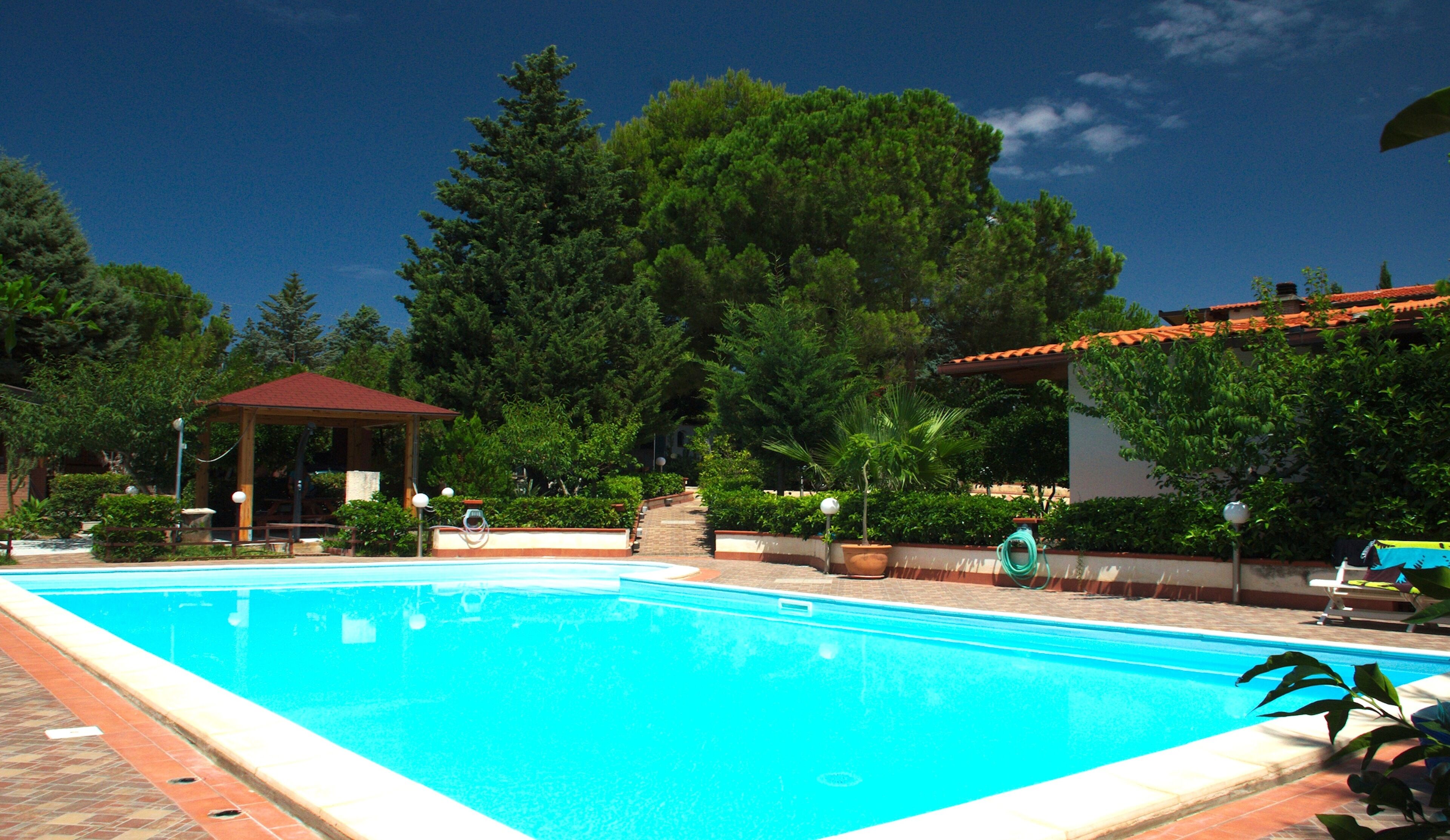 Ein Pool mit einem pavilIon mit rotem Dach, umgeben von üppiger Vegetation und wolkenlosem, blauem Himmel.