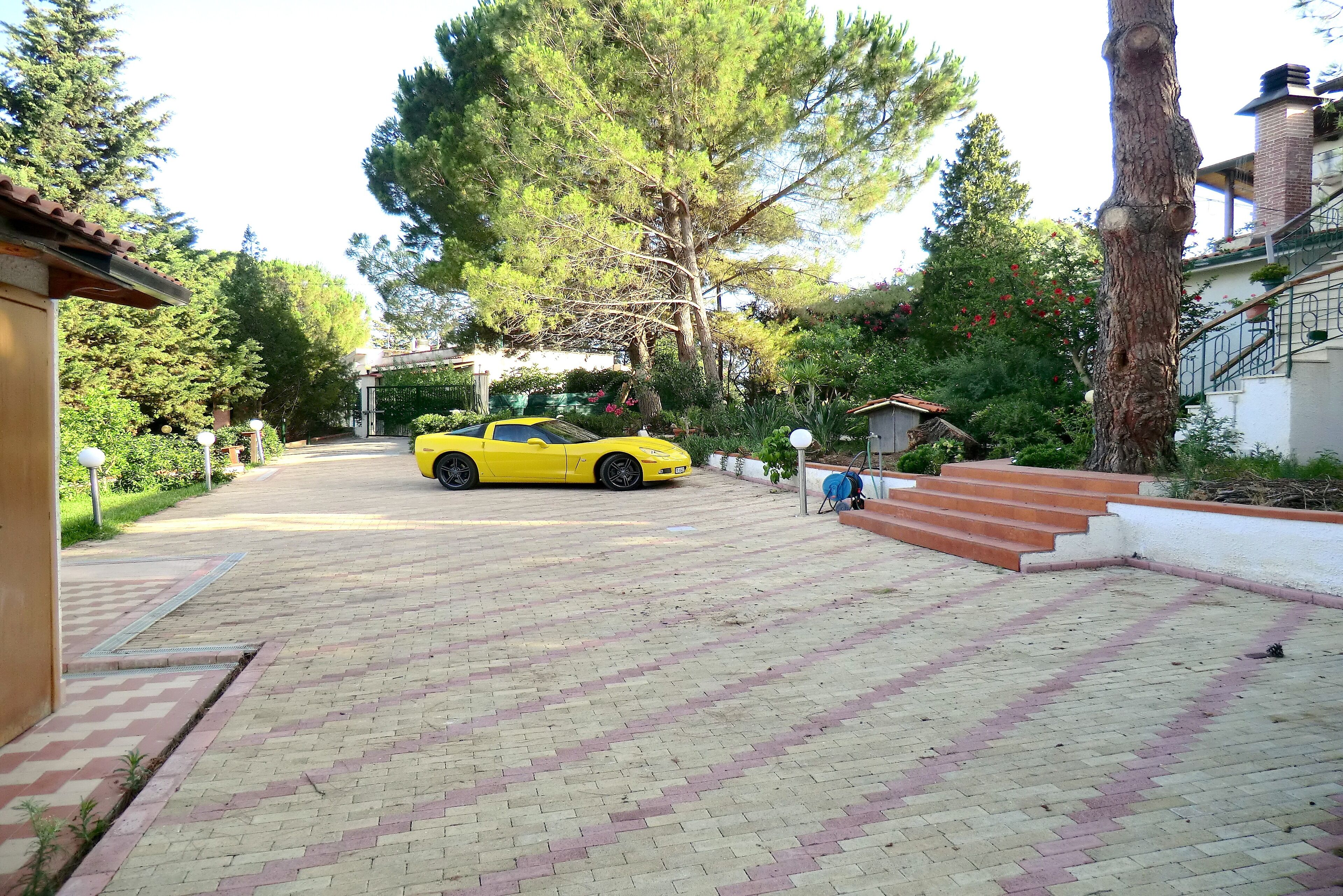 A yellow car parked on a paved driveway with a brick pattern, surrounded by trees and a white building with a red roof.