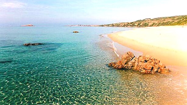 Ein Strand mit klarem türkisblauem Wasser, einem sandigen Ufer und einem Felsen.