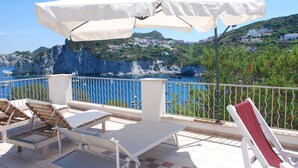A terrace with sun loungers and a white umbrella, overlooking a coastal area with cliffs and houses. - The house by the sea one step away from the beach (ponza)