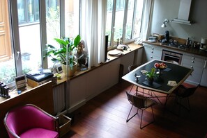 A kitchen with a dining table, chairs, and a red chair. There is a window with curtains, a plant, and a small statue on the counter.
