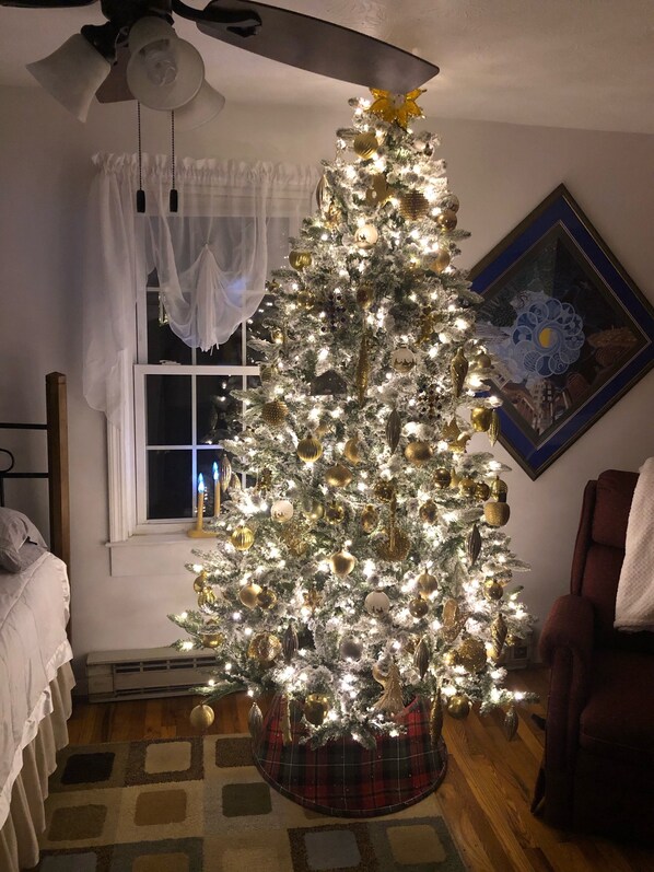 A room with a Christmas tree decorated with lights and ornaments, a ceiling fan, a window with curtains, a framed picture on the wall, and a red chair.