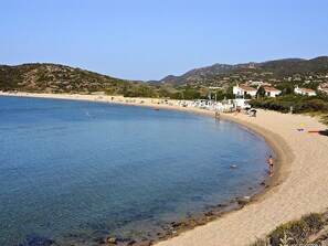 Ein Strand mit klarem, blauem Wasser, sandigem Ufer und Gebäuden im Hintergrund.
