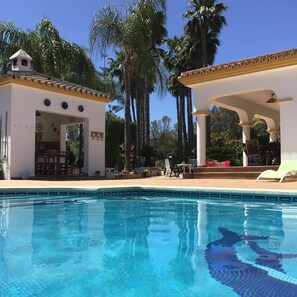 A swimming pool with clear blue water, surrounded by palm trees and covered outdoor seating areas.