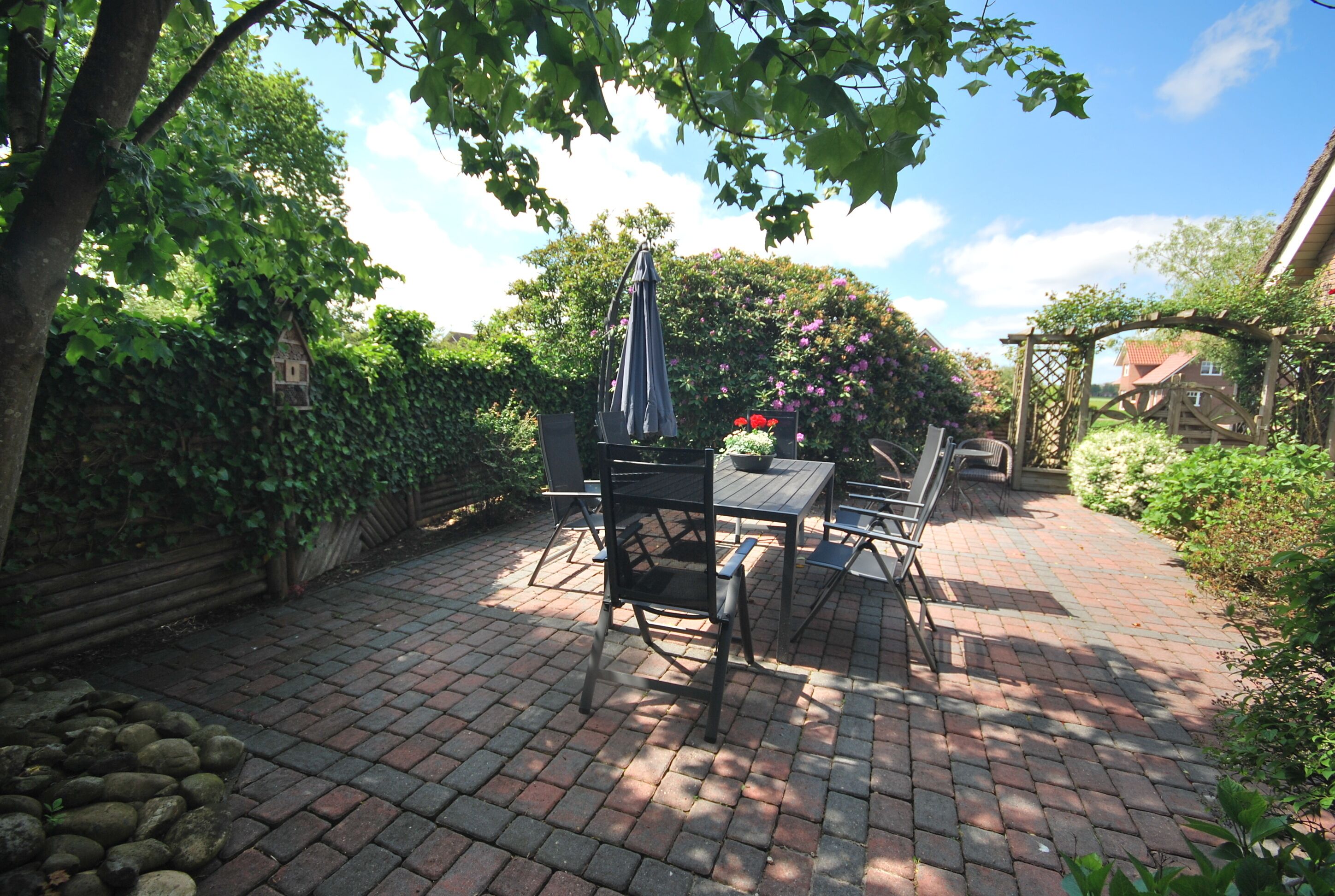Une terrasse en briques avec une table et des chaises, entourée de verdure et d’une clôture en bois.