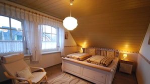 A bedroom with a wooden ceiling, a bed with a headboard, a rocking chair, a window with curtains, and a hanging light fixture. - 4 * holiday on the canal in Nordgeorgsfehn (East Frisia) (Uplengen)