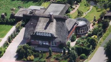 Una vista aerea di una grande casa storica con una torre circolare imponente, circondata da giardini curati e un vialetto lastricato.