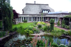 Une grande maison à toit de chaume, entourée d’un jardin bien entretenu avec un étang et diverses plantes.