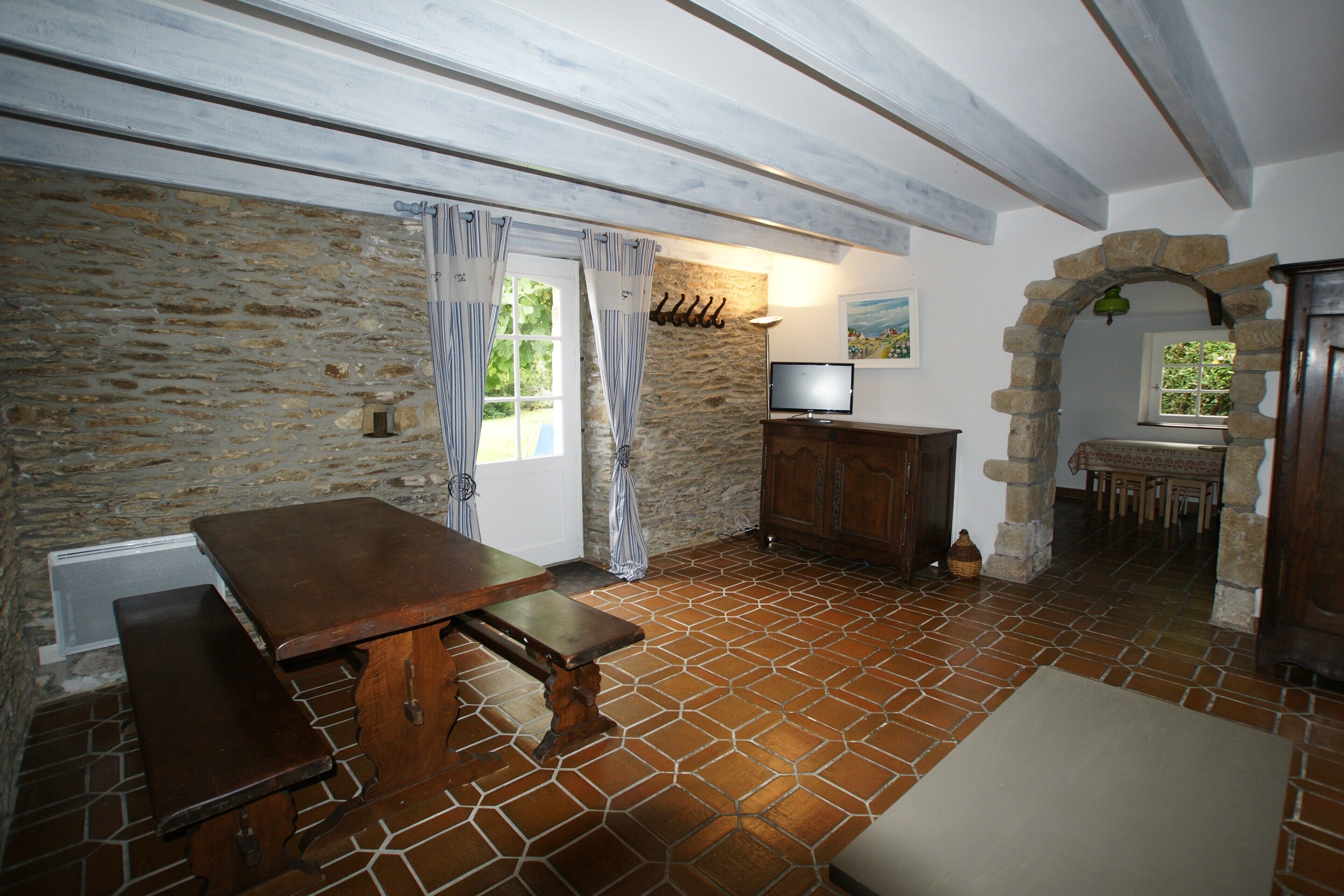 Une pièce rustique avec un mur en pierre, une table en bois avec des bancs, un téléviseur et une arche en pierre.