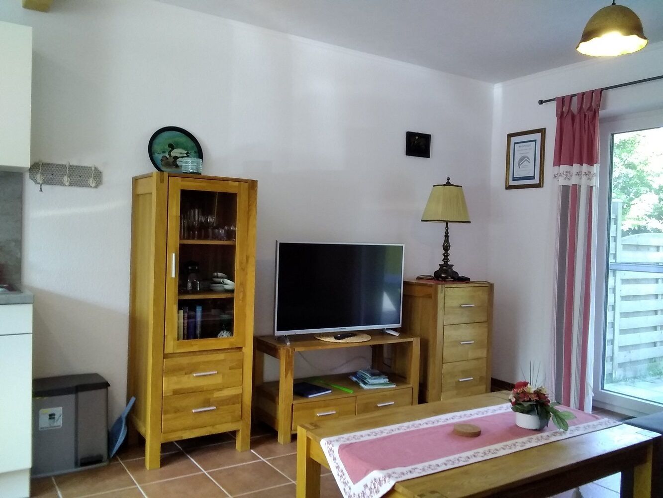 Ein kleiner, gemütlicher Wohnraum mit einem Holzkabinett, einem Fernseher auf einem Schrank, einem kleinen Tisch mit einer Matte und einem Fenster mit Vorhängen.