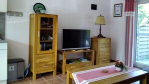 A small, cozy living room with a wooden cabinet, a TV on a stand, a small table with a mat, and a window with curtains.
