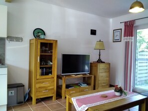 Uma sala de estar pequena e acolhedora, com um armário de madeira, uma TV sobre um móvel, uma mesa com um tapete e uma janela com cortinas.