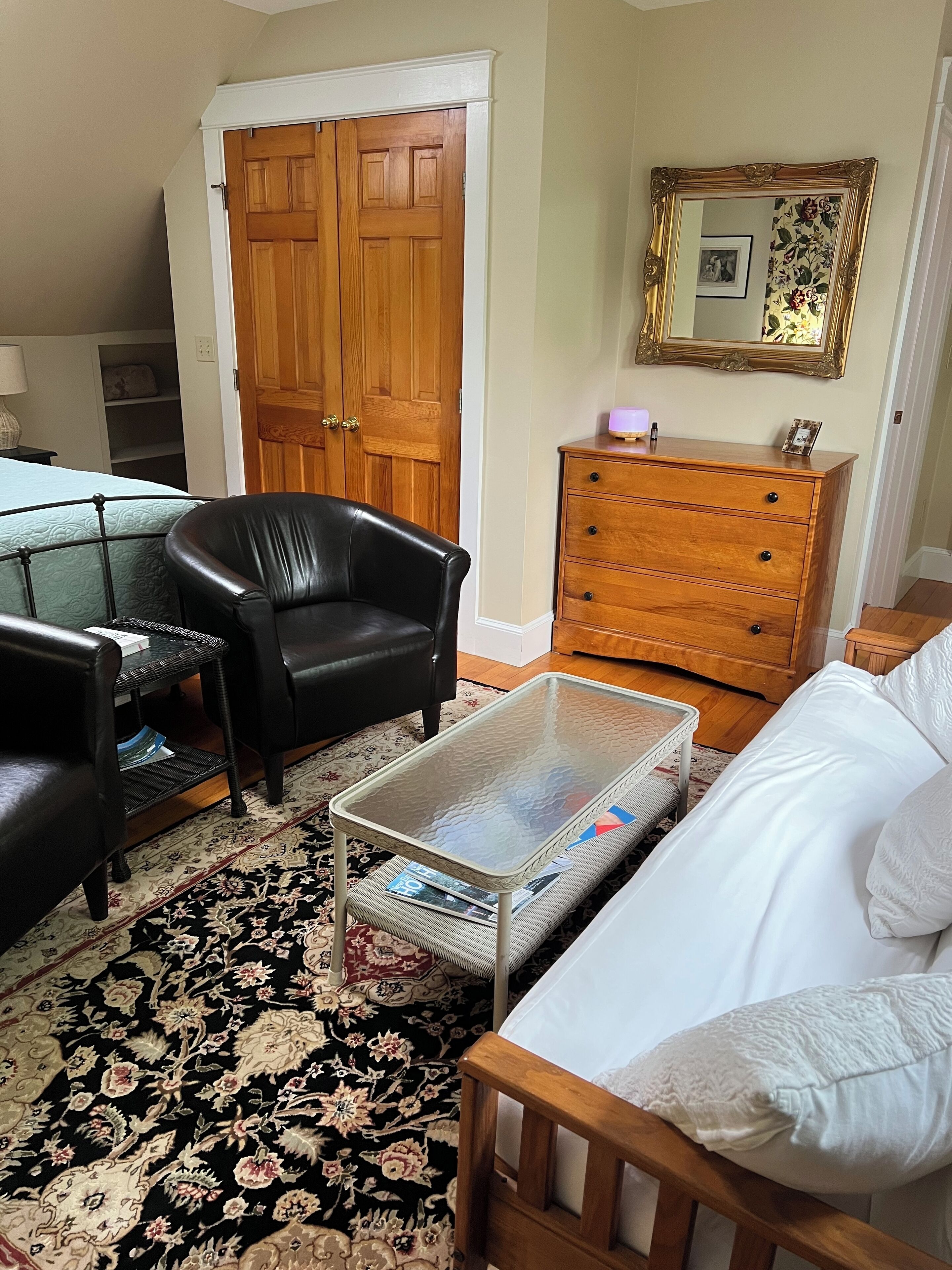 Een kamer met een zwarte leren fauteuil, een glazen salontafel met tijdschriften, een witte bank, een houten dressoir en een houten deur.