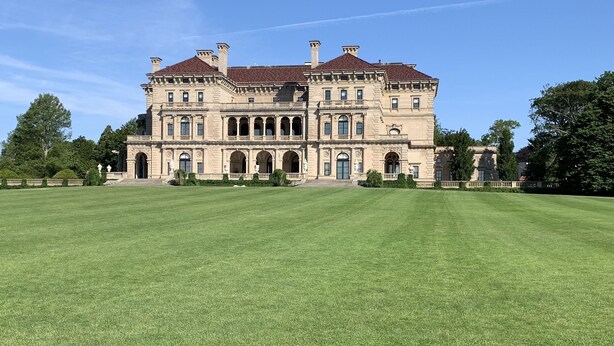 A large, historic building with a symmetrical facade, multiple arches, and a spacious lawn in front.
