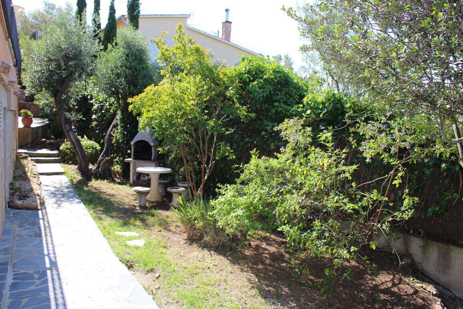 A garden with a stone table and chairs, a small stone structure, and various shrubs and trees.