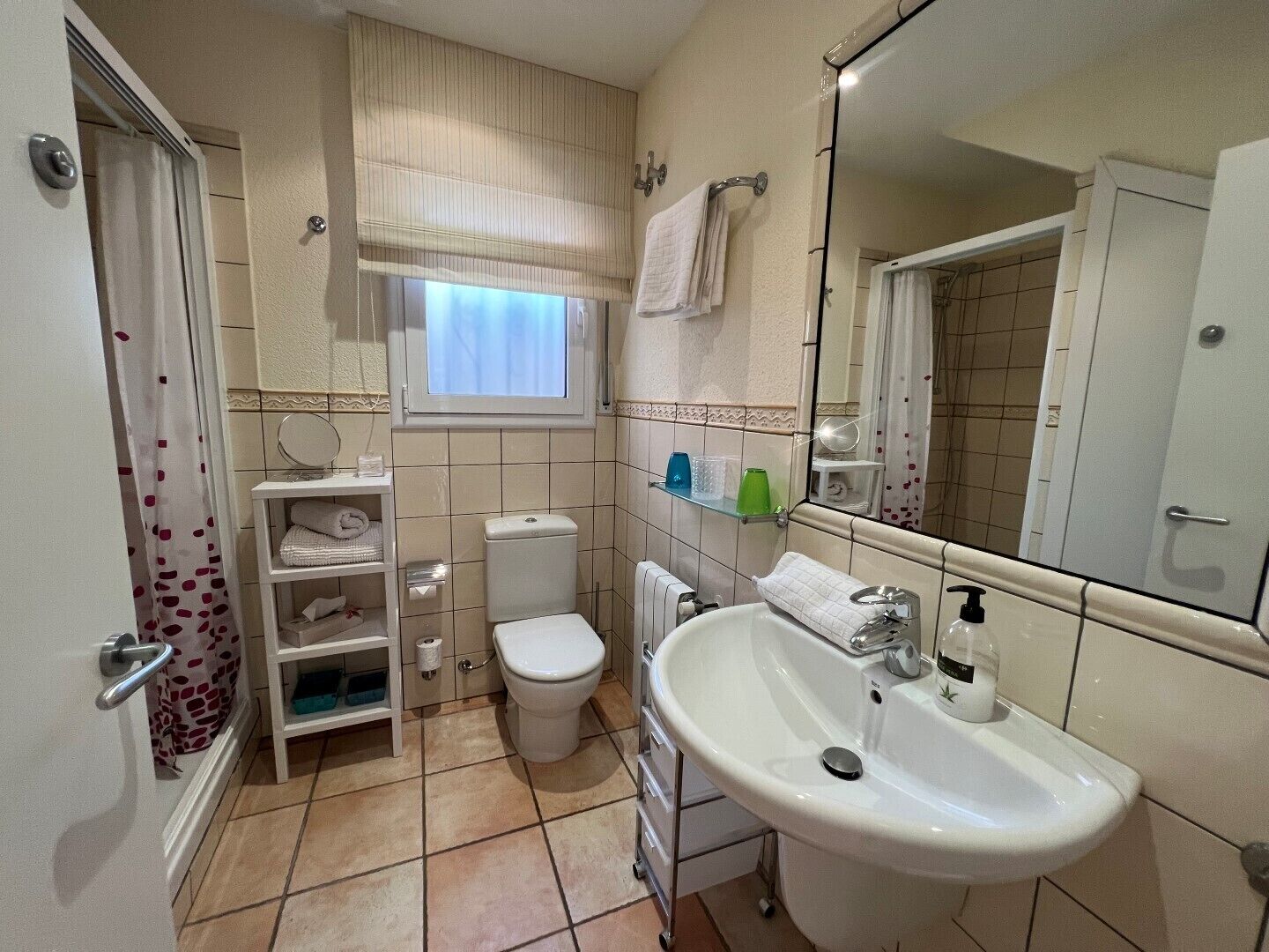 Een badkamer met toilet, wastafel en spiegel. Er is een raam met gordijn, een handdoekhouder en planken met toiletartikelen.