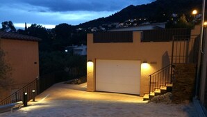 Une allée pavée de pierres mène à un garage avec une porte blanche, qui est entouré d’un petit muret en pierre et d’un escalier doté d’une rampe en métal.