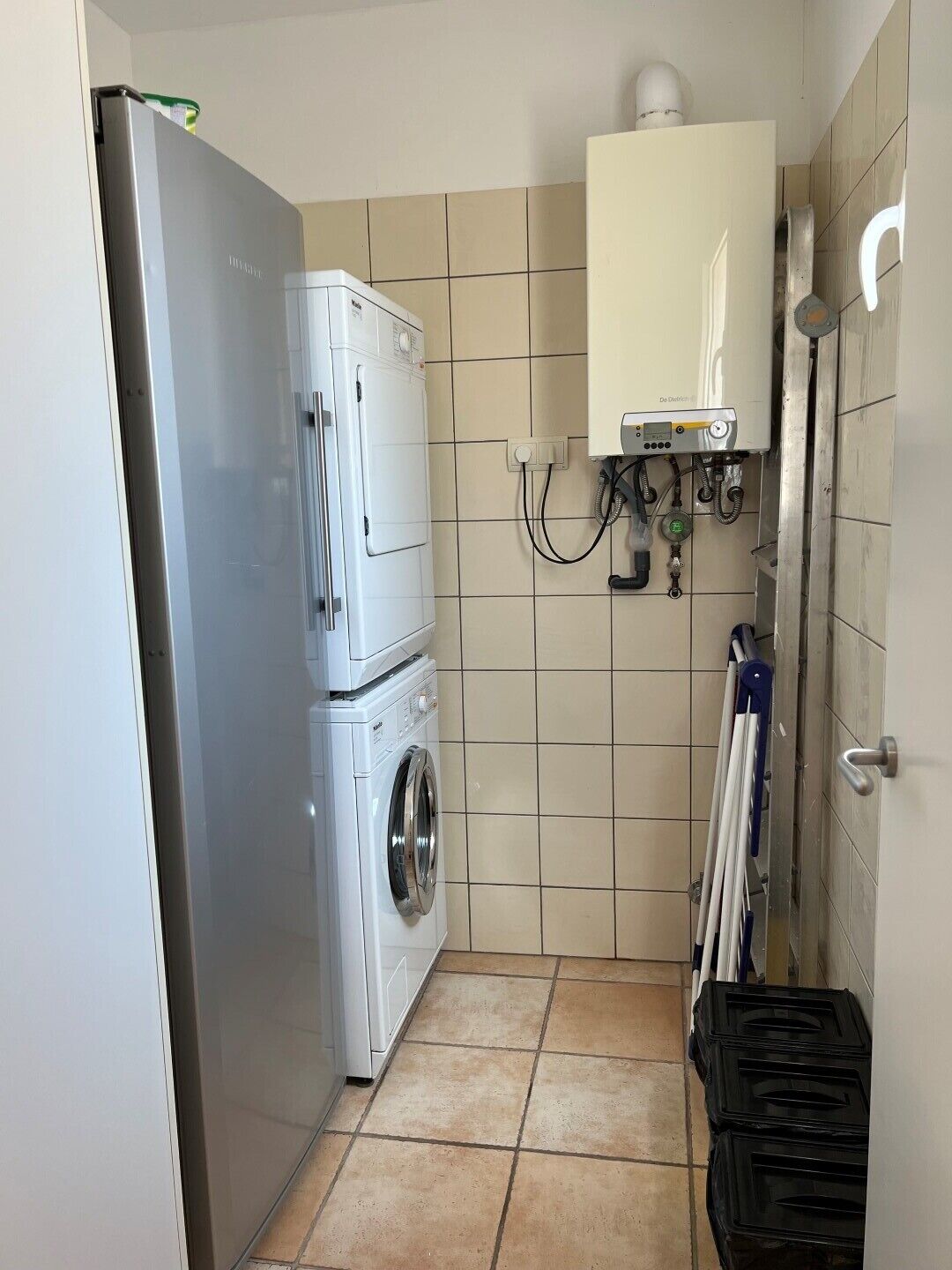 Une petite buanderie équipée d’un lave-linge et d’un sèche-linge superposés, d’un chauffe-eau et d’une étagère en métal où sont rangés les produits de nettoyage.