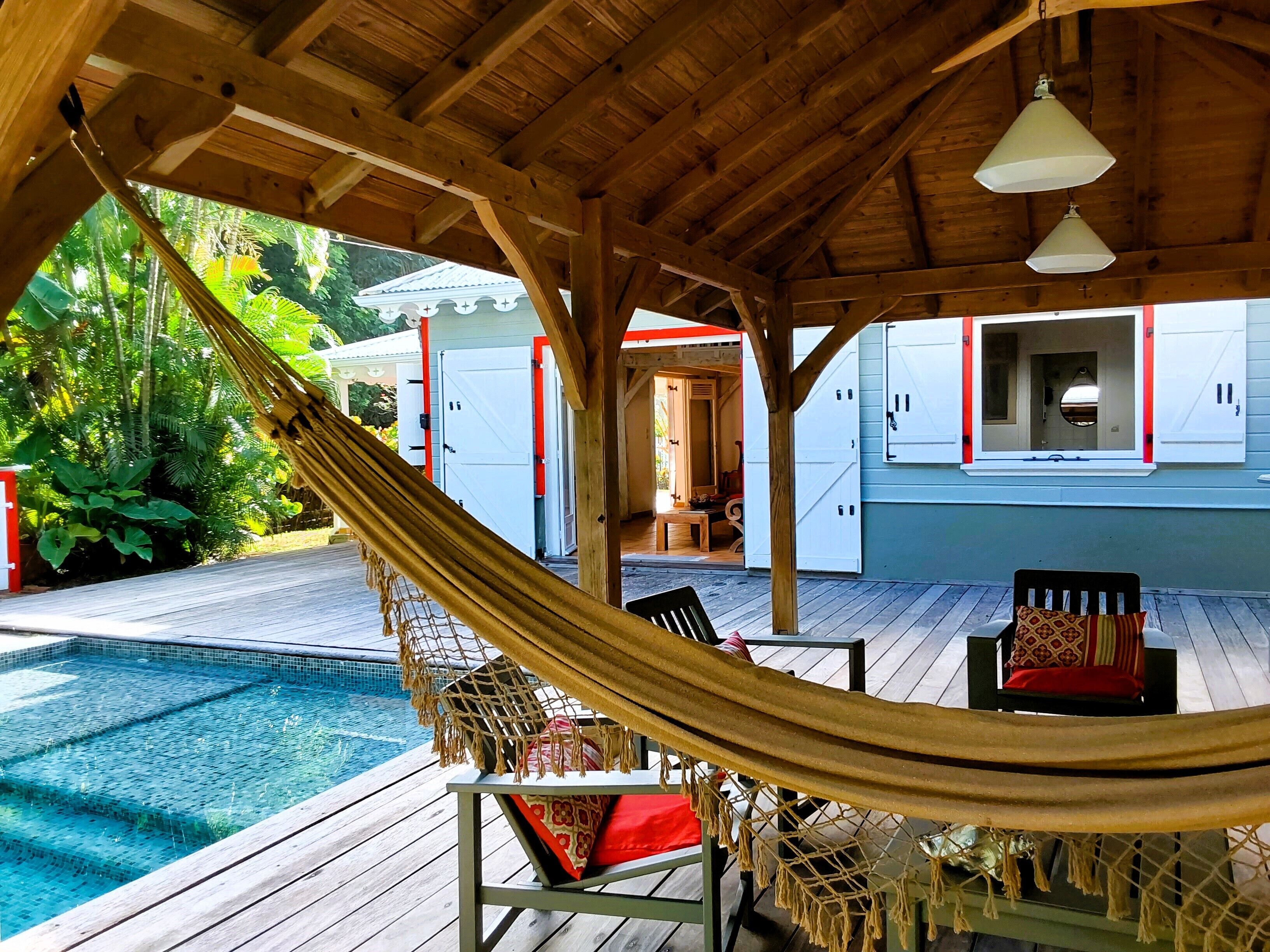 A wooden deck with a hammock, a pool, and a building with white walls and red trim.