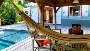 A wooden deck with a hammock, a pool, and a building with white walls and red trim. - Villa Emeraude - Superb Creole villa with amazing Pool & direct access to beach (Diamant)
