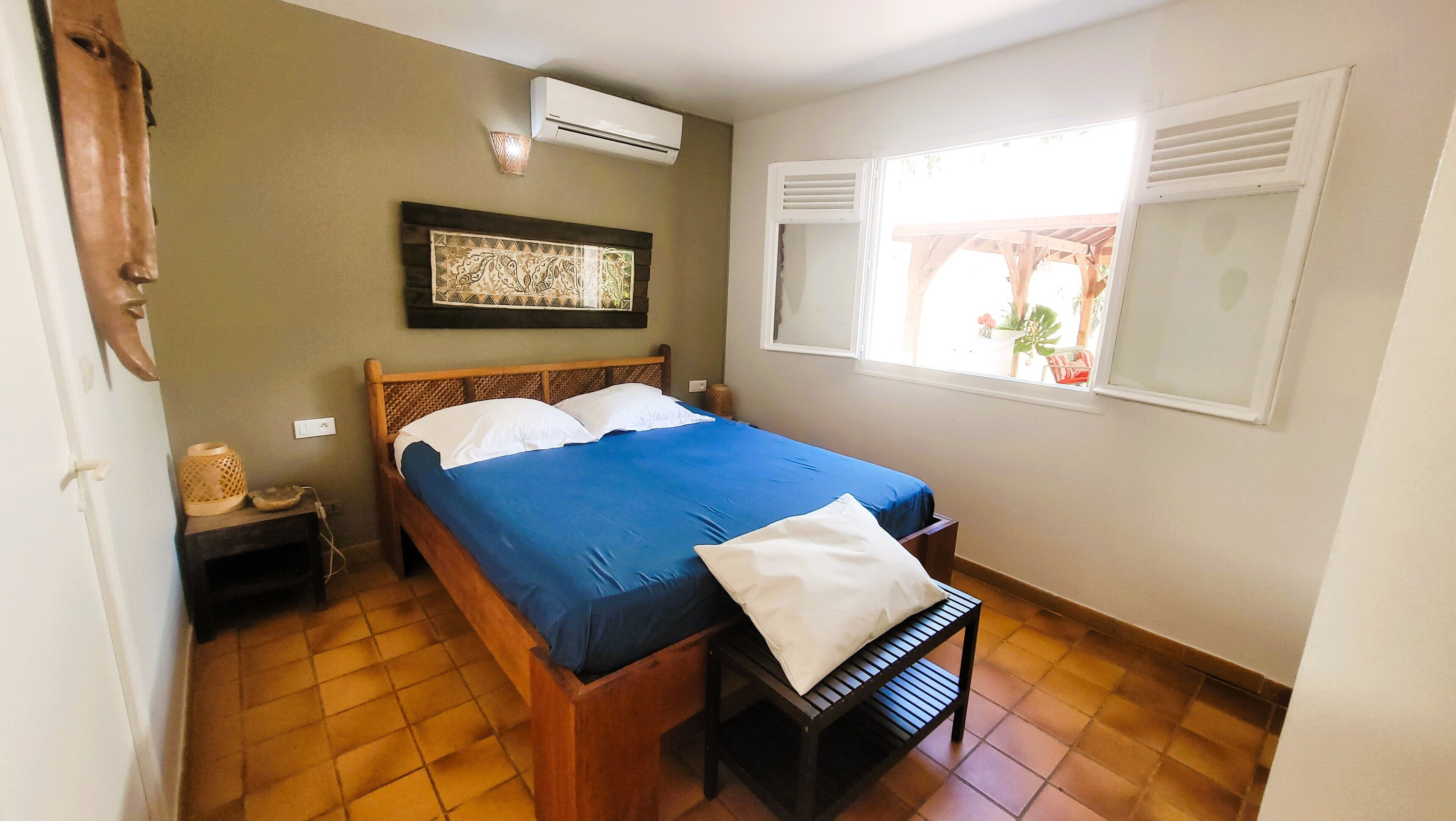 Une chambre à coucher comprenant un lit, une table de chevet, un climatiseur, une fenêtre avec des volets et une œuvre d’art accrochée au mur.