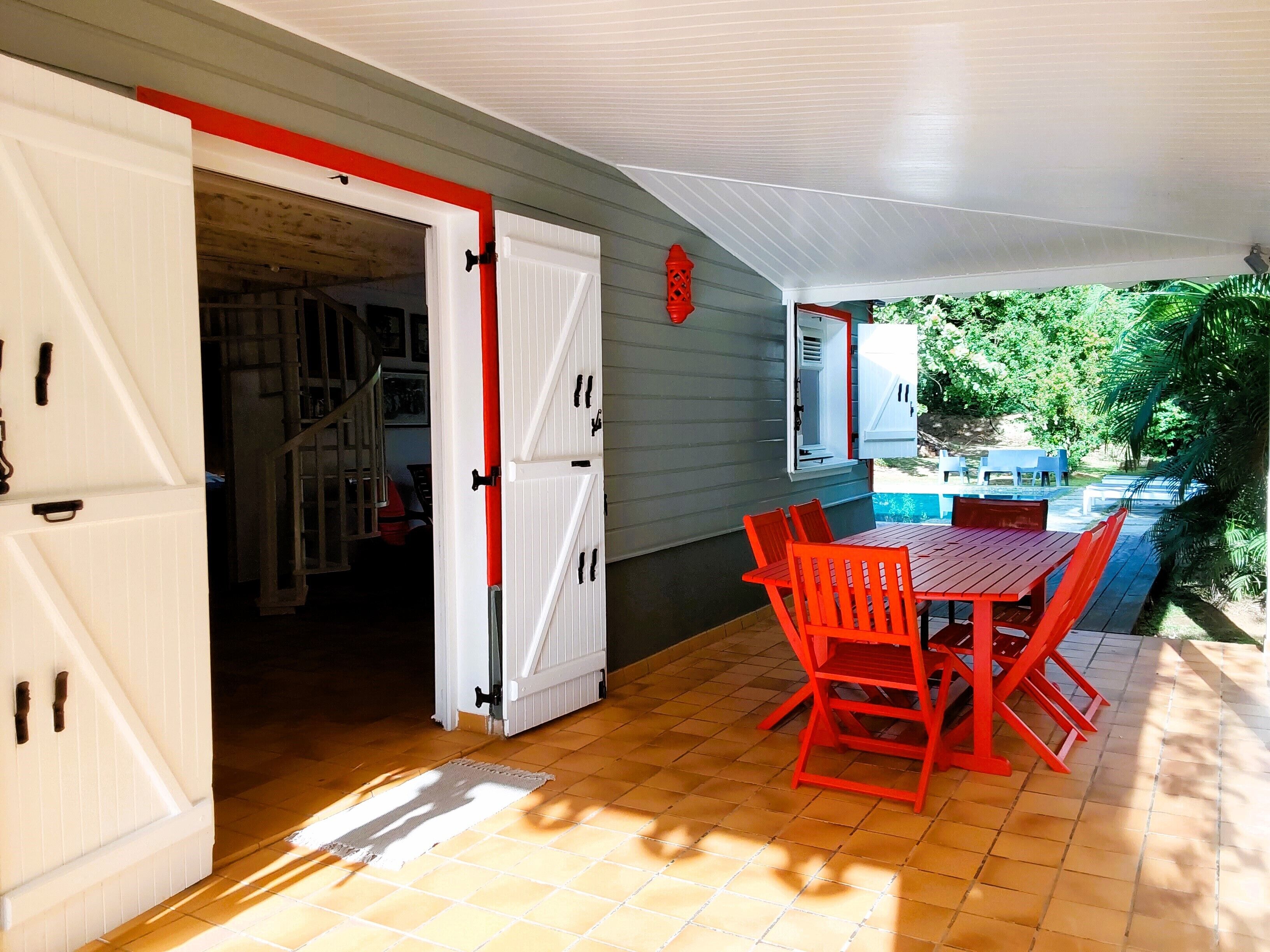 A covered patio with a red table and chairs, a white door, and a view of a pool and greenery.