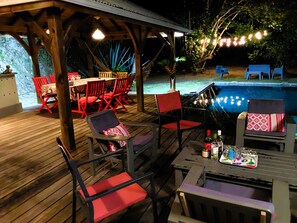 An outdoor patio with wooden flooring, red and black chairs, a table with drinks, and a pool in the background.