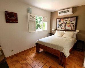 A bedroom with a bed, a window with shutters, an air conditioner, and a painting on the wall.