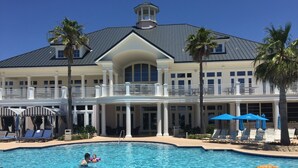 A large swimming pool with lounge chairs and umbrellas, surrounded by a two-story building with a balcony.