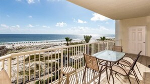 A balcony with a glass table and chairs, overlooking a beach with palm trees and a clear sky.