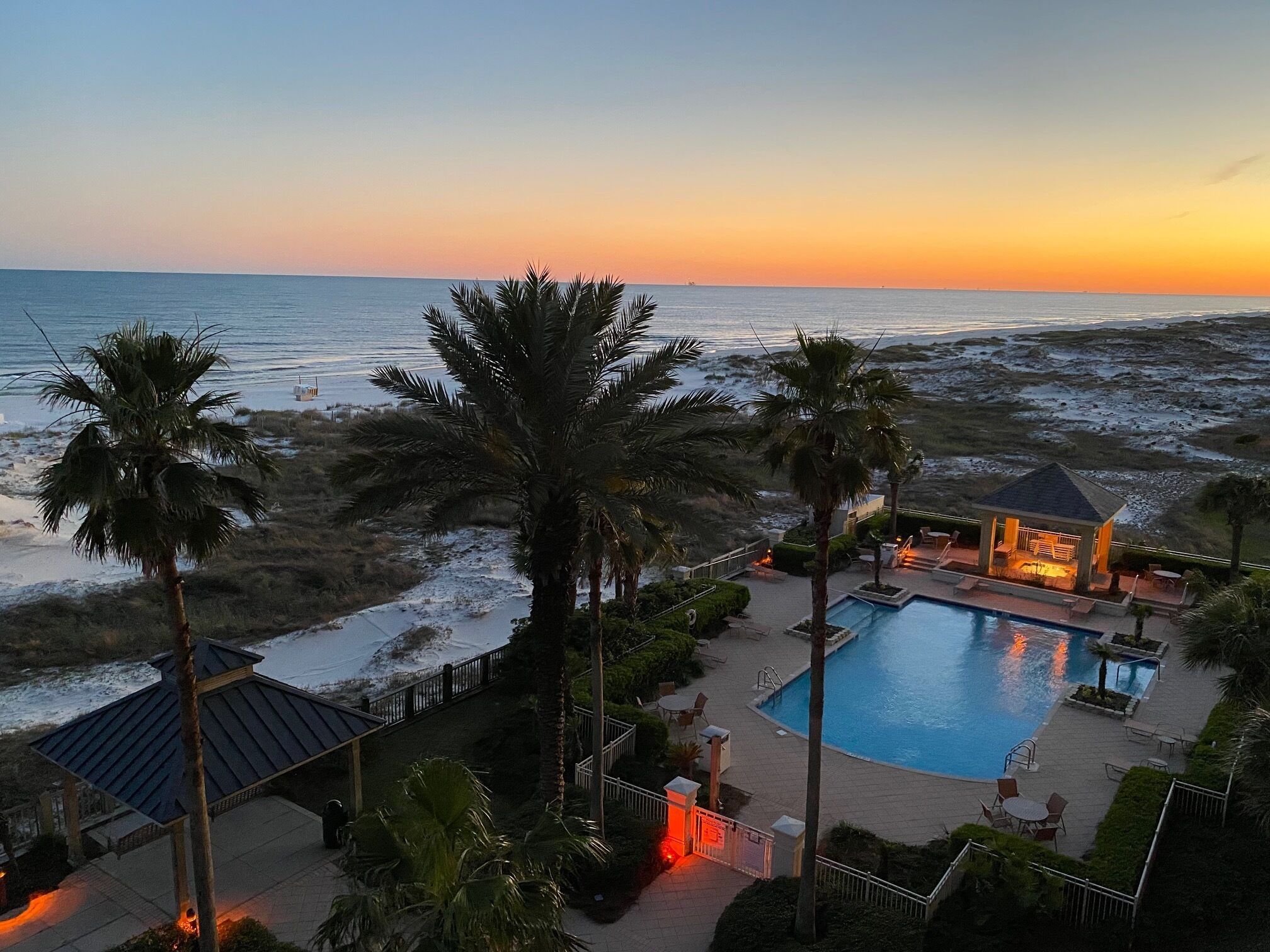 A resort with a swimming pool, palm trees, and a view of the ocean at sunset.