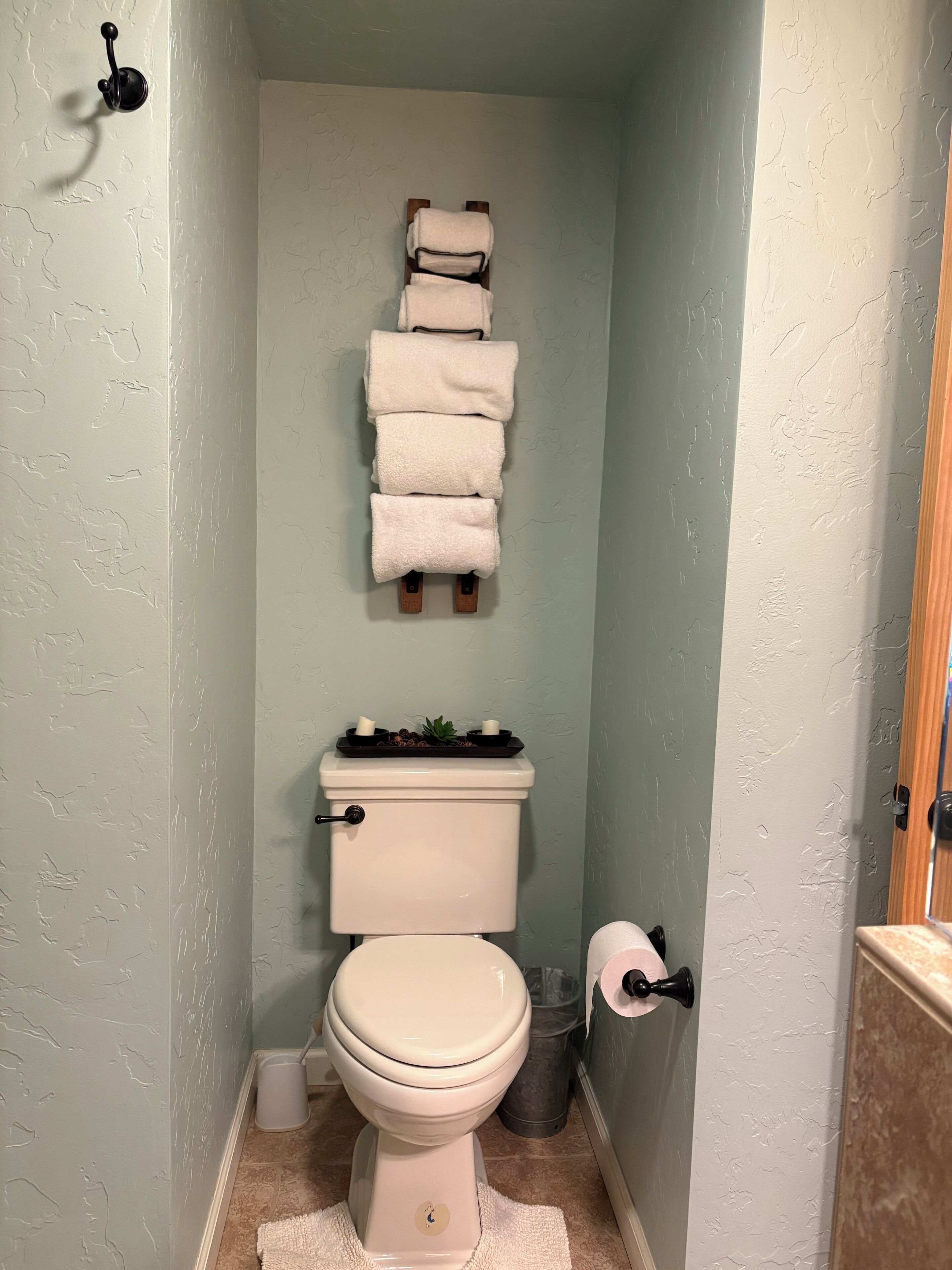 Un baño con inodoro, una pila de toallas, un estante pequeño con artículos de aseo y un cesto de basura.