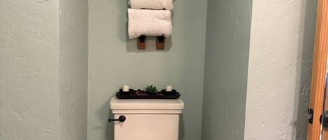 Un baño con inodoro, una pila de toallas, un estante pequeño con artículos de aseo y un cesto de basura.