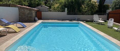 Una piscina rectangular con agua azul cristalina, rodeada por una terraza de madera y una zona de césped con sillas de descanso.