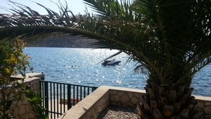 A stone wall with a palm tree, a railing, and a view of a calm sea with a boat.
