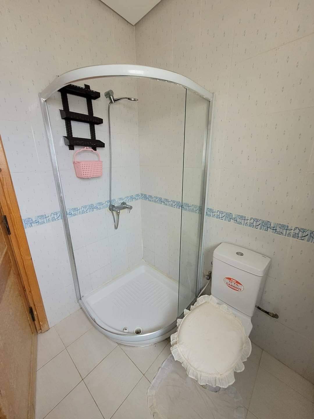 Un petit espace douche fermé, doté d’une porte en verre, de toilettes blanches et d’un petit panier rose fixé au mur.