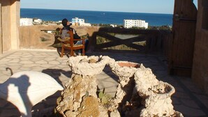 A stone patio with a wooden chair and a person sitting. A large, open gate leads to a view of the sea and buildings in the background. - Vacation apartment in Tunisia (quiet location) (CHATT MERIAM)