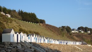 Eine Reihe von Strandkörben mit einer Treppe, die zu ihnen hinaufführt, auf einem sandigen Strand mit einem felsigen Vordergrund und einer Hügelkette mit Vegetation und Häusern im Hintergrund.