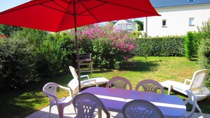 Eine Terrasse mit Tisch und Stühlen, einem roten Sonnenschirm und einem Garten mit rosa Blumen.