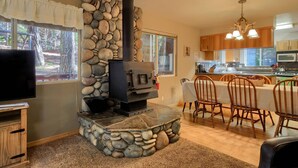 Une cheminée en pierre avec un téléviseur posé sur un meuble en bois, un coin repas avec des chaises et une table, et une cuisine avec des armoires en bois et un lustre.