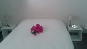 Una cama bien hecha con un ramo de flores rosadas en el centro, flanqueado por dos mesitas de noche con lámparas y un teléfono.