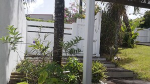 Un jardín con cerca de piquetes blancos, diversas plantas y un camino de concreto que lleva a una puerta blanca.