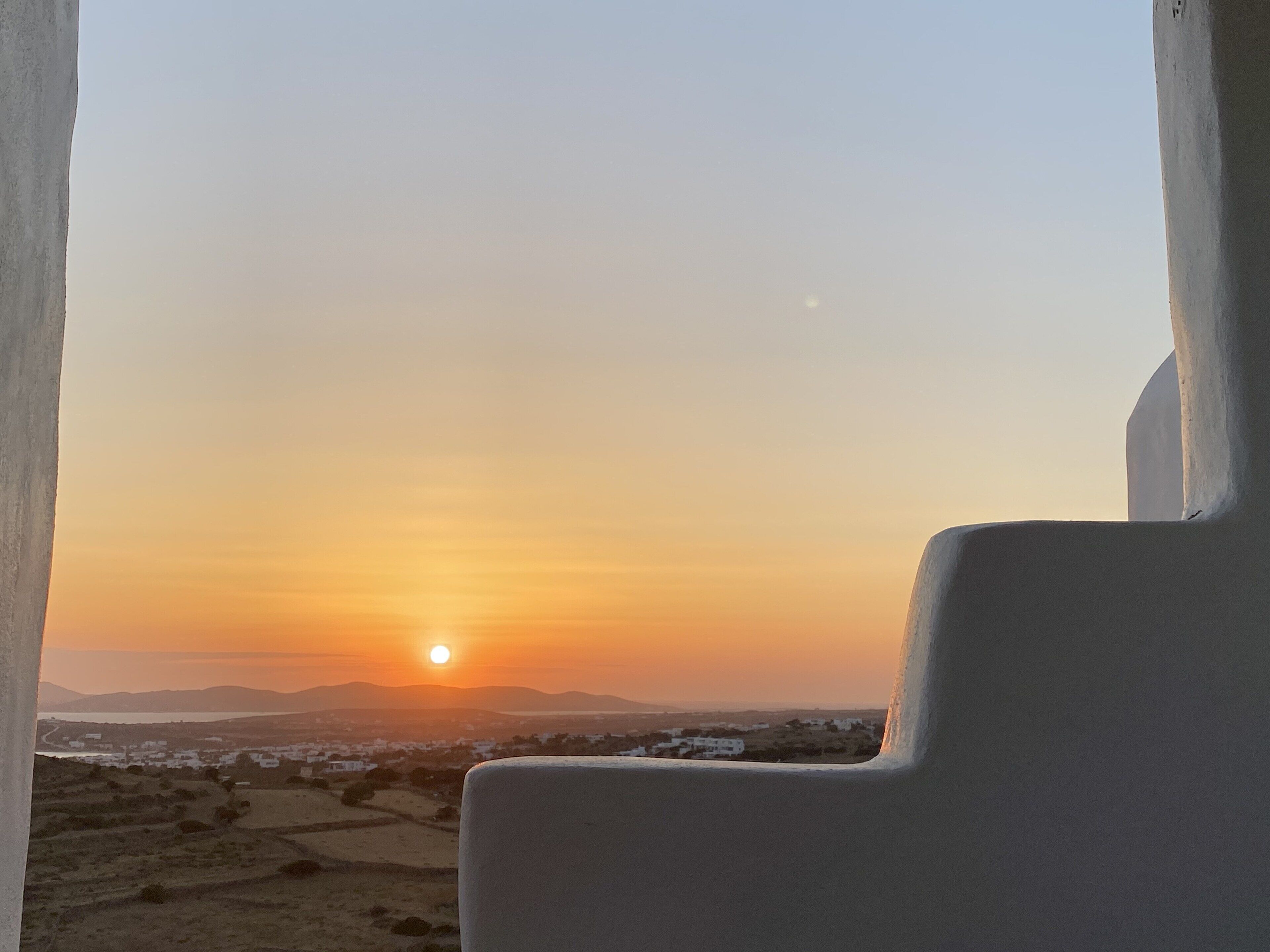 A view from a window overlooking a sunset over a landscape with distant mountains and a few buildings.