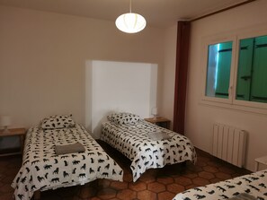 A room with two beds, each with patterned bedding, a bedside table with a lamp, a window with green curtains, and a radiator. - LARGE VILLA 12 PERS. 5 mn from the beaches. LARGE SWIMMING POOL AND 2000m2 LAND. (Six-Fours-Les-Plages)