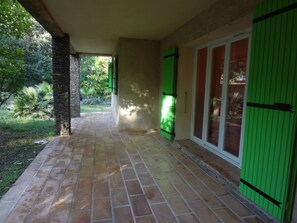 A covered patio area with a brick floor, green shutters, and a view of the surrounding greenery. - LARGE VILLA 12 PERS. 5 mn from the beaches. LARGE SWIMMING POOL AND 2000m2 LAND. (Six-Fours-Les-Plages)