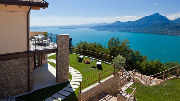 Une maison moderne avec une façade en pierre, un balcon agrémenté de mobilier d’extérieur et une vue sur un lac et des montagnes.