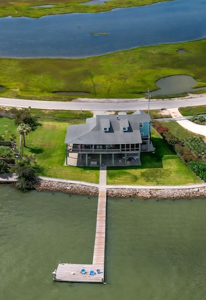 An aerial view of a modern house with a dock extending into a body of water, surrounded by grassy areas and a small pond. - 4 3.5 Bayside 2 King Masters, A+ Sunset, Pier Fishing Light,Huge Cover Porch (Galveston)