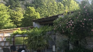 A rustic outdoor seating area with wooden loungers and a stone building surrounded by lush greenery and a mountainous backdrop. - Into the wild: Charming house, surrounded by nature, large private pool. (Romeyer)